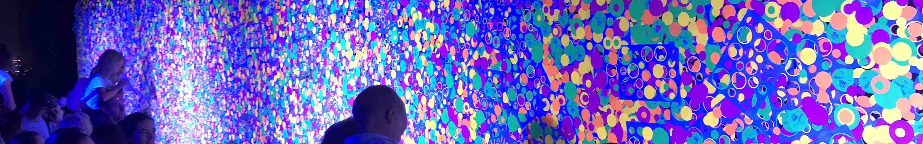 Crowd at public event with wall of glow-in-the-dark dots illuminated by ultraviolet light.