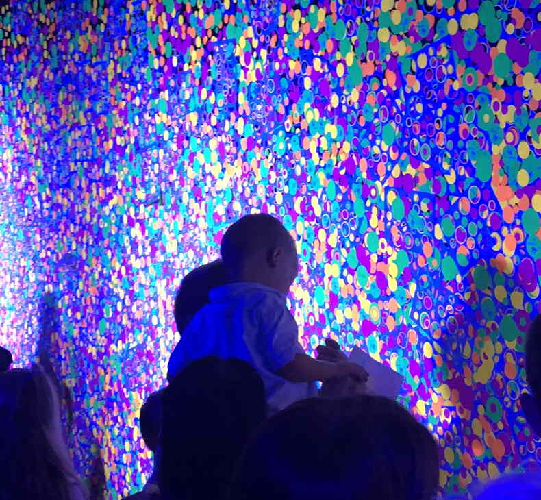 Crowd at public event with wall of glow-in-the-dark dots illuminated by ultraviolet light.